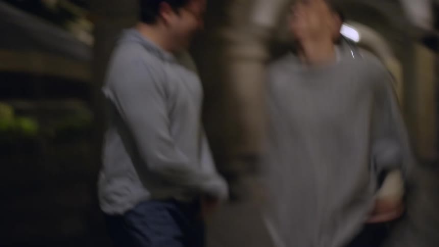 A young man and woman laugh and smile while walking together through a dimly lit outdoor stone corridor with arched ceilings at night