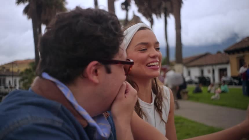 A man with glasses and a woman with a white headband sit together outdoors and laugh while looking at a smartphone screen in a park with tall palm trees
