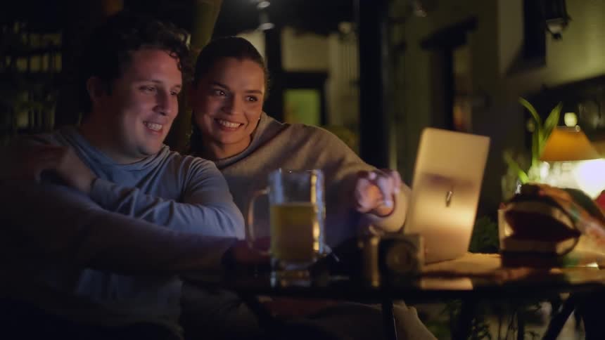 In this short clip a young man and woman are sitting together outdoors at night illuminated by the glow of a laptop screen they are smiling and looking at the computer while a large glass of beer sits on the table in front of them