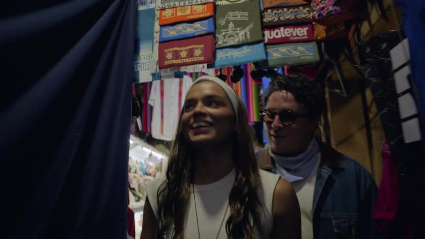 Woman and man walk through a market with colorful stalls and hanging products
