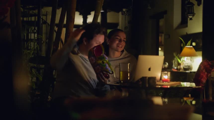 Couple seated at a table in a nighttime setting using a laptop and smartphone with drinks and candle lighting around