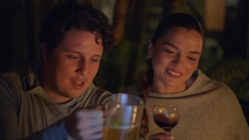 A man with curly hair and a woman smile while holding up a large mug of beer and a glass of red wine respectively during an evening gathering outdoors