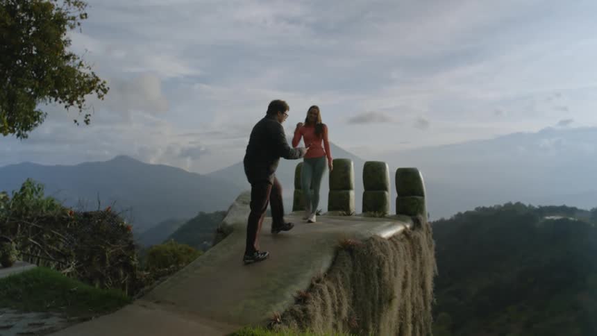 Person photographs a woman with arms outstretched at a high viewpoint overlooking mountains