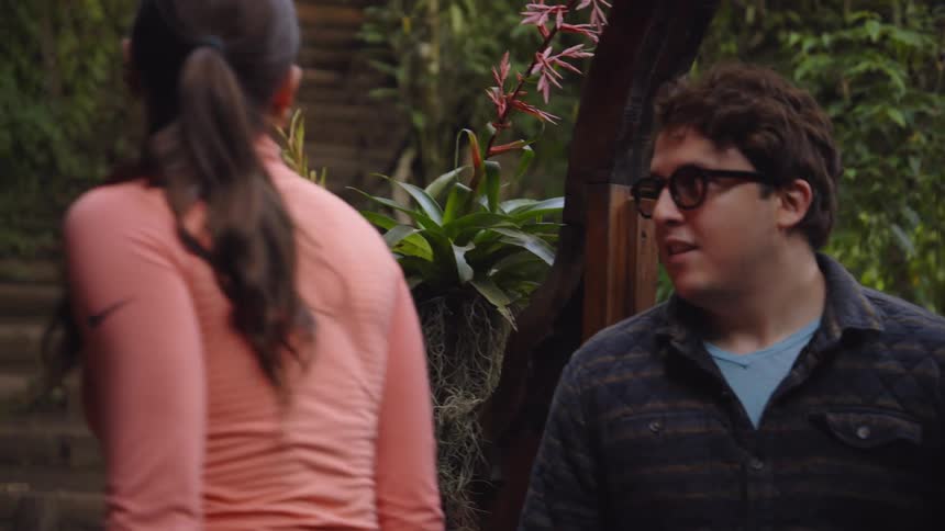 A man and a woman walk together up a stone path lined with lush greenery they chat as they transition from a narrow walkway toward a set of stairs leading deeper into the garden