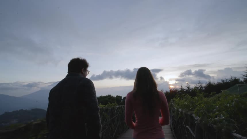 A woman in a pink top and a man in a jacket walk along a wooden path toward a mountain sunset while the woman briefly holds up her phone to capture the view of the clouds and peaks