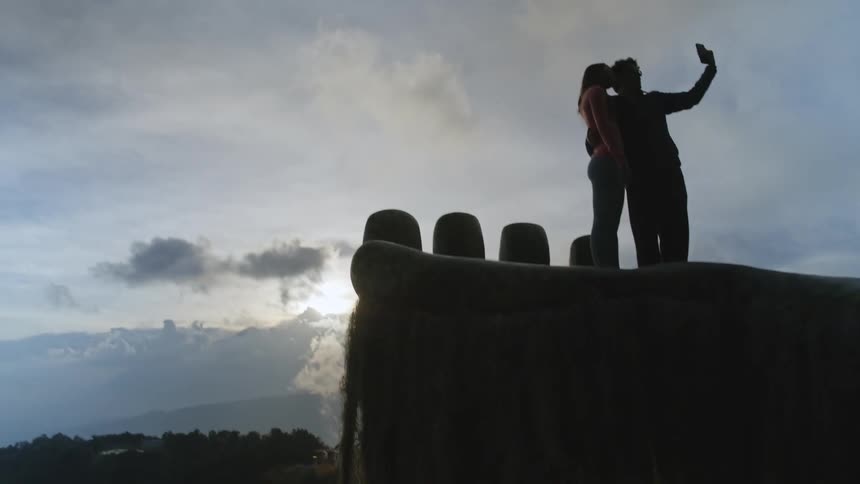 A couple stands on a large handshaped sculpture overlooking a misty mountain valley during sunset they are silhouetted against the bright sky as they hold up a phone to take a selfie together