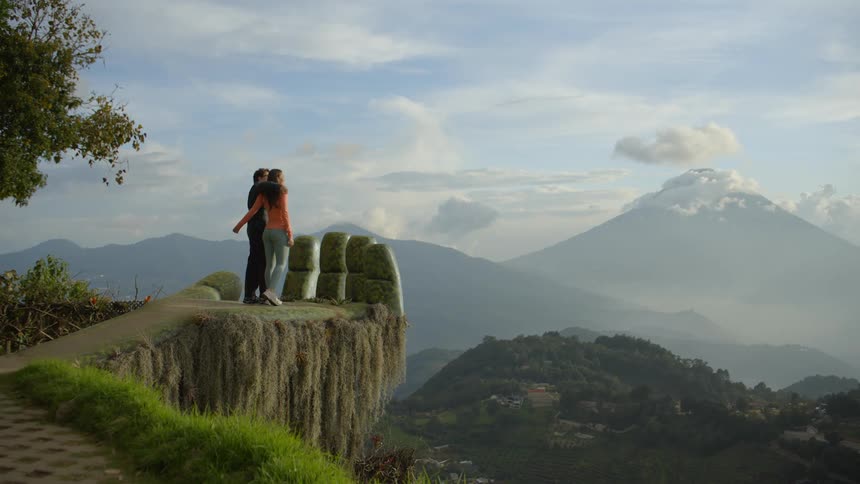 A couple holding hands walking on the statue palm at hobbitenango ecopark watching the wide natural horizon at midday