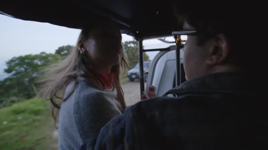 A woman with long hair and a man wearing glasses sit in the back of an openair vehicle as it drives along a winding mountain road