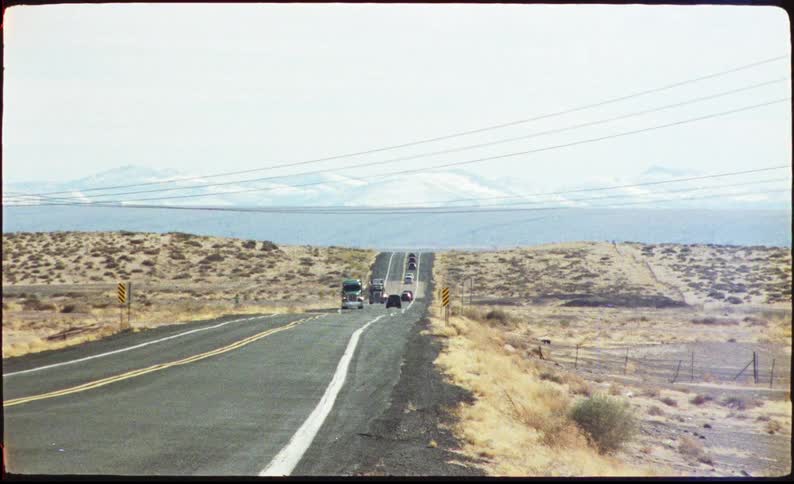 A series of cars and trucks driving along a countryside road at midday with vast open land stretching out on both sides