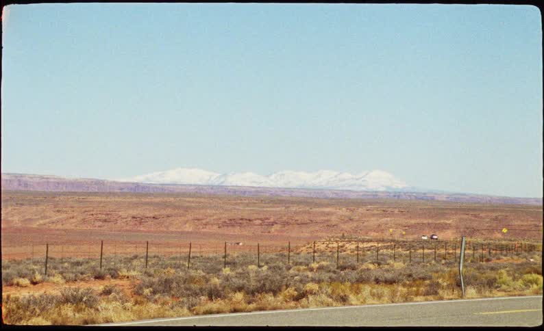 The video captures a wide desert landscape featuring red rock mesas and distant snowcapped mountains under a clear sky it is filmed with a vintage aesthetic characterized by a visible film border and a slight grain