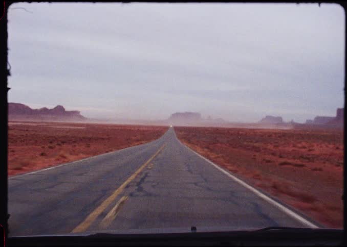 A vehicle drives down a long straight highway cutting through a vast desert landscape with reddishbrown terrain large rock formations and mesas are visible in the distance under a hazy overcast sky