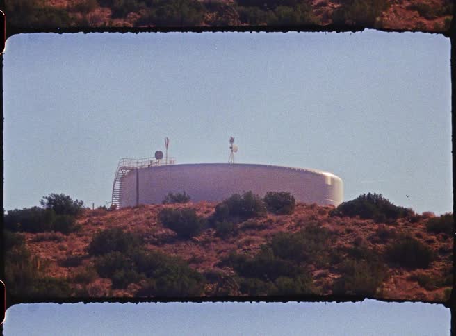 The video features a large white industrial storage tank situated on top of a brushcovered hill the footage has a vintage grainy aesthetic with a filmstrip border suggesting a retro or cinematic style