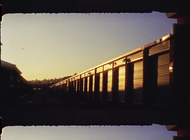 A lowangle view shows a long row of outdoor storage units with closed metal rollup doors illuminated by the warm orange light of a setting sun