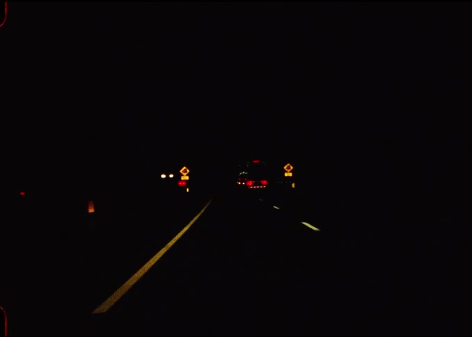 A camera mounted inside a vehicle captures a firstperson perspective of driving down a dark highway at night passing illuminated yellow road signs and following the red taillights of another car while white lane markers recede into the distance