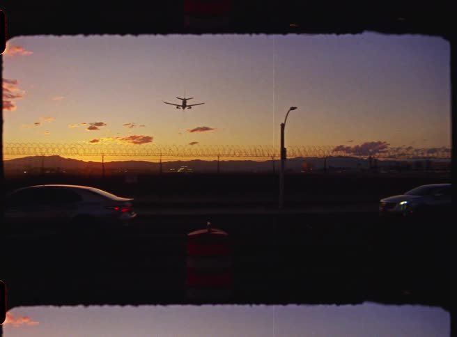 Airplane flies over a busy road and towards an airport as red tints the sky above the mountains