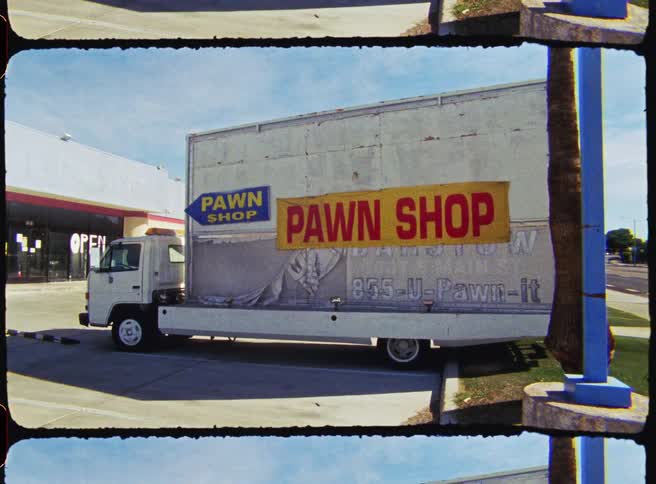 A truck with the word pawn shop in large capital letters printed on a bright yellow rectangular banner that is attached to the side of a truck