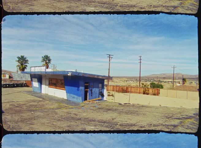 A dilapidated blue and white building sits abandoned in a cracked asphalt lot under a wide desert sky