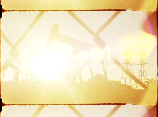 Oil pumpjacks seen through a fence at sunset with power transmission towers in the background