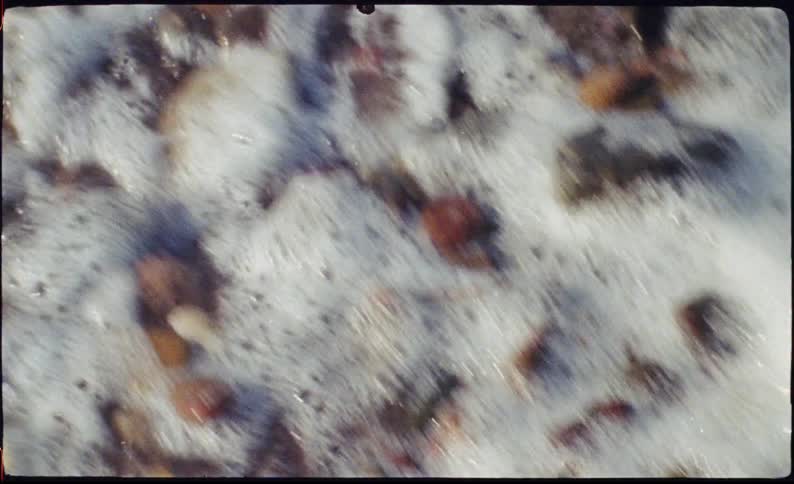 This video shows a closeup topdown view of ocean waves gently washing over a pebbly shore the footage has a vintage grainy aesthetic that mimics the look of old film