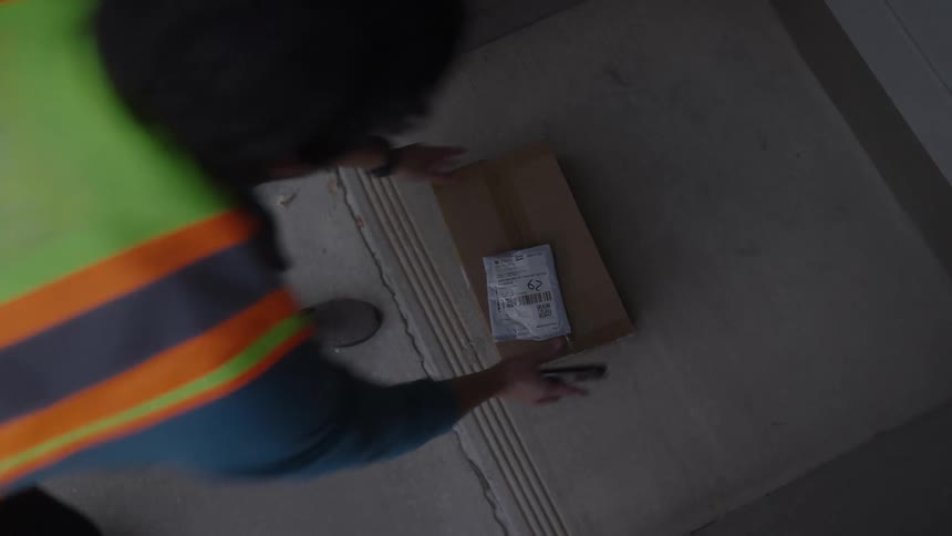 A delivery worker in a safety vest places a cardboard package on a concrete door step
