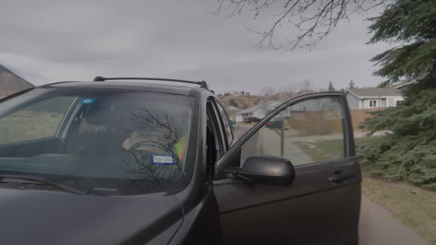 A young man wearing a green vest opening his car door at midday carrying a box as he steps out of a car parked on the side of the street
