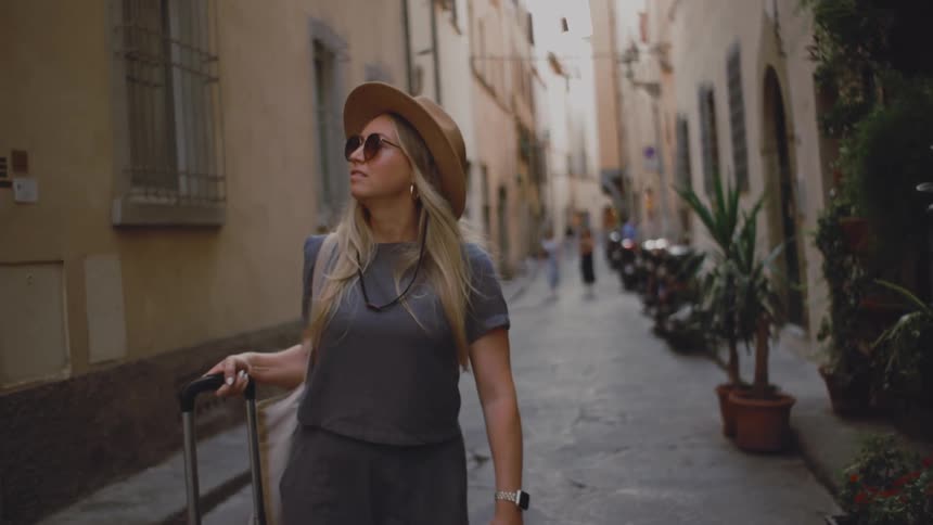 Woman walking down a narrow street pulling a rolling suitcase wearing a hat and sunglasses with potted plants and old buildings around