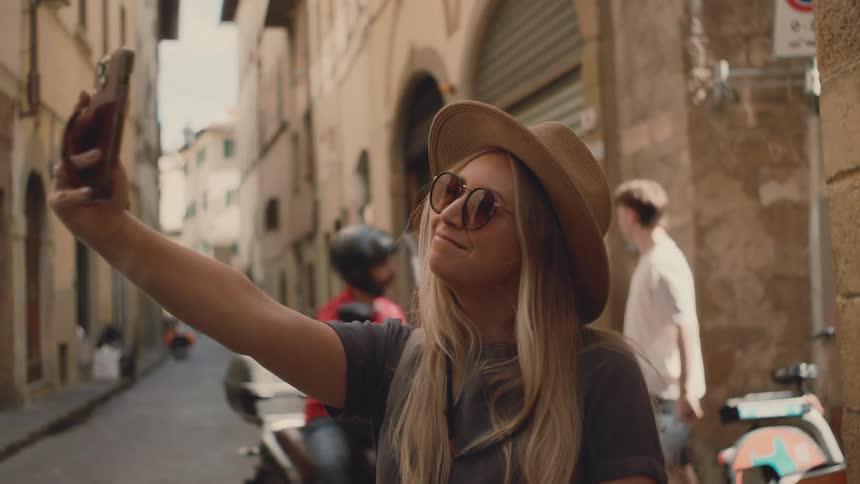 Woman holds a smartphone to take a selfie on a narrow street with pedestrians in the background