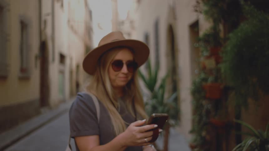 Woman walking along a narrow street pulling a rolling suitcase wearing a hat and sunglasses surrounded by potted plants and old buildings