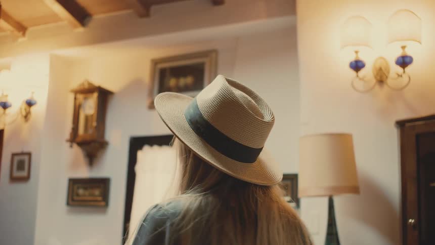A young woman walking through a building smiling as she looks at the furniture while wearing a sunhat