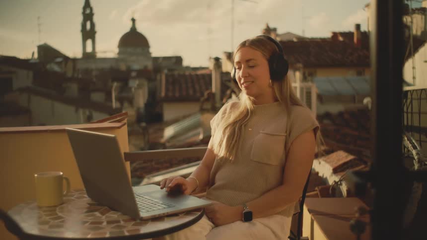 Woman uses a laptop with headphones on a balcony overlooking the city at sunset