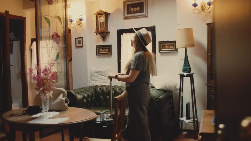 A woman wearing a widebrimmed hat walks into a vintagestyle room while carrying a suitcase she pauses in the center of the space looking around with a smile at the antique furniture and decor