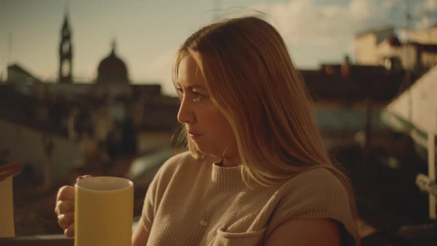 Woman drinks from a mug on a balcony overlooking the city at sunset