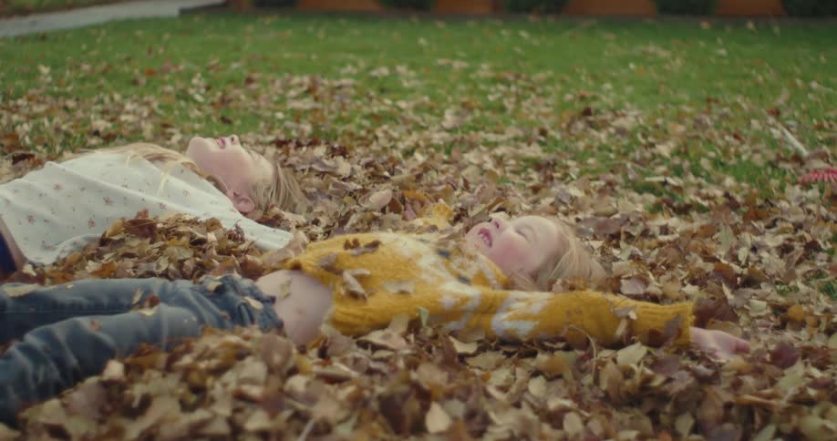 Two young girls sitting in the front yard fall backward into the leaves as they play