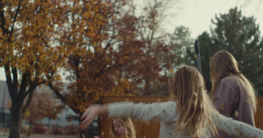 Two young girls playing in the front yard and throwing leaves into the air as their mother rakes the leaves
