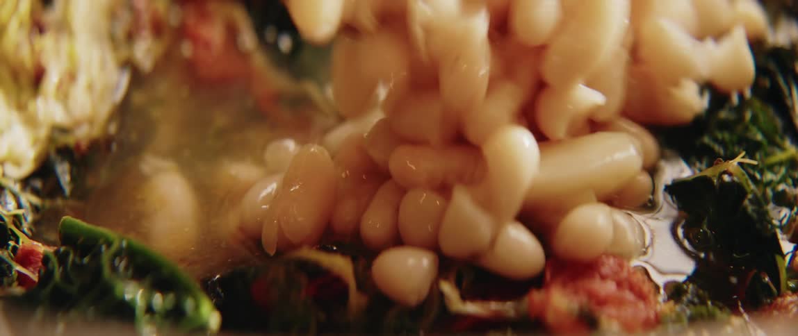 An extreme closeup shot shows a large spoonful of white beans being poured into a steaming pot containing cooked greens and tomatoes
