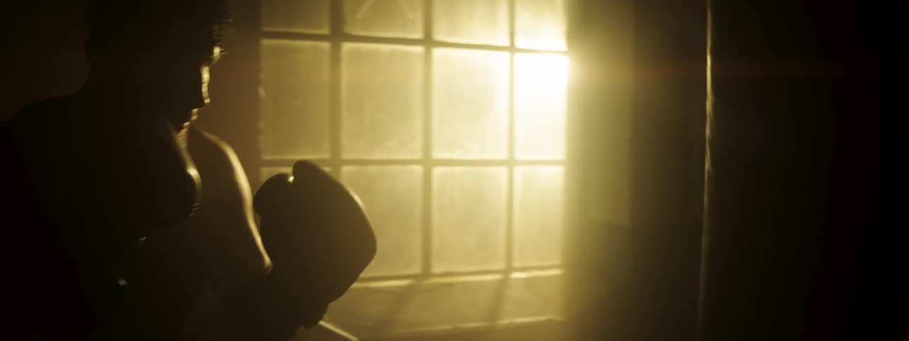A male boxer in silhouette repeatedly strikes a heavy bag with his gloves inside a dimly lit gym as golden sunlight streams through a large window in the background