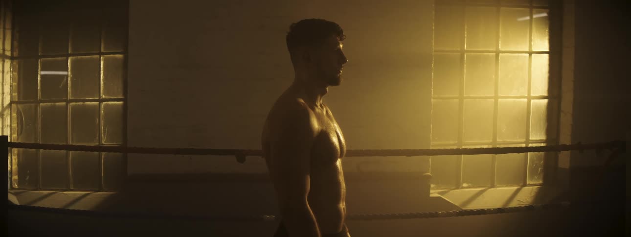 A side view of a shirtless man standing in a boxing ring as warm light streams through a window into a dark gym