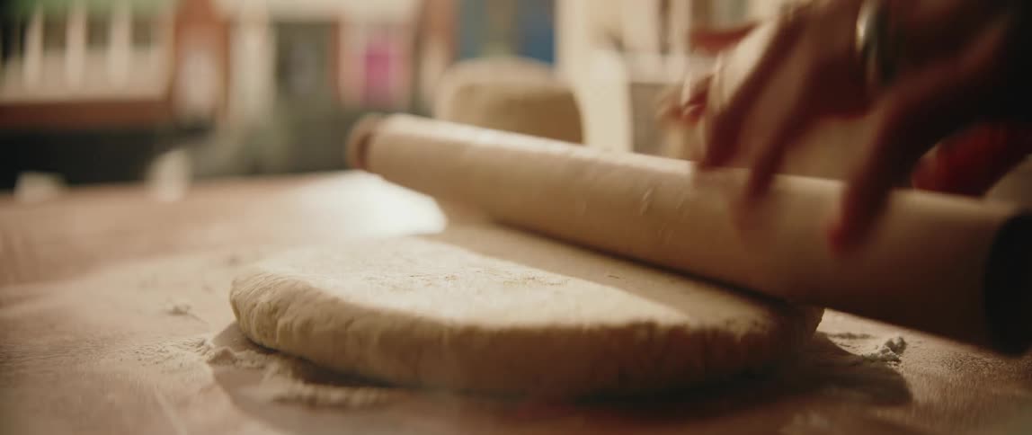 A close view shows a persons hands using a wooden rolling pin to flatten dough on a floured surface then sprinkling more flour over the top and spreading it with their hand
