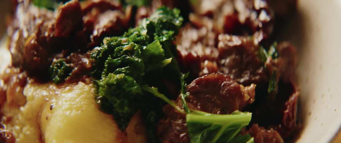 A closeup camera pan shows a dish of tender braised beef chunks served over creamy mashed potatoes and vegetables