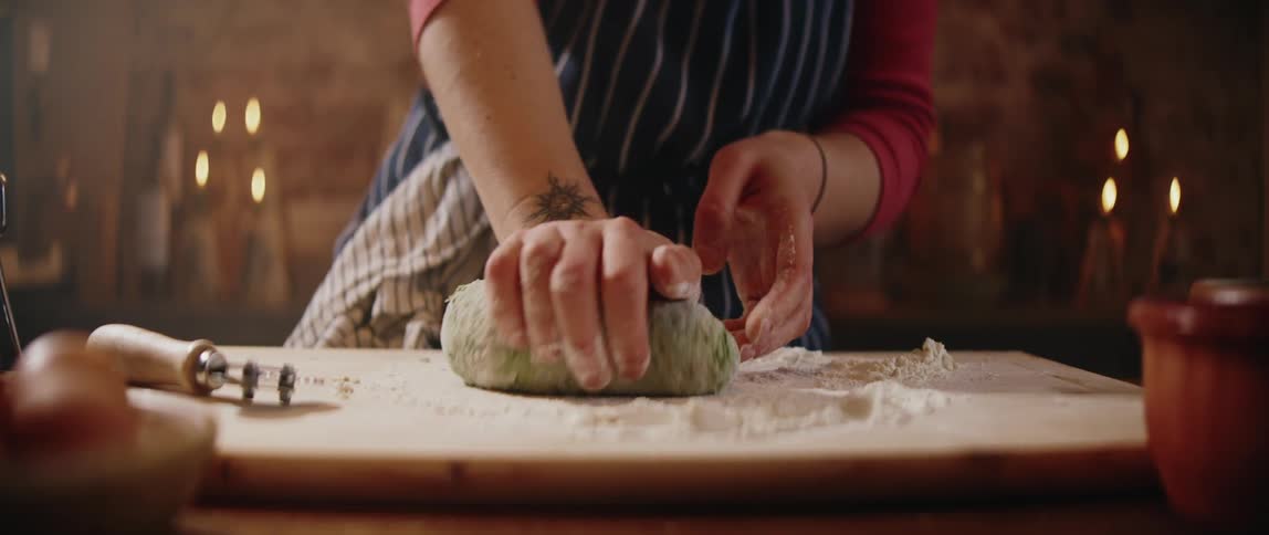 A woman in a striped apron and red shirt kneads a ball of green herbinfused dough on a floured wooden board using her hands to press and fold the mixture in a rustic dimly lit kitchen setting