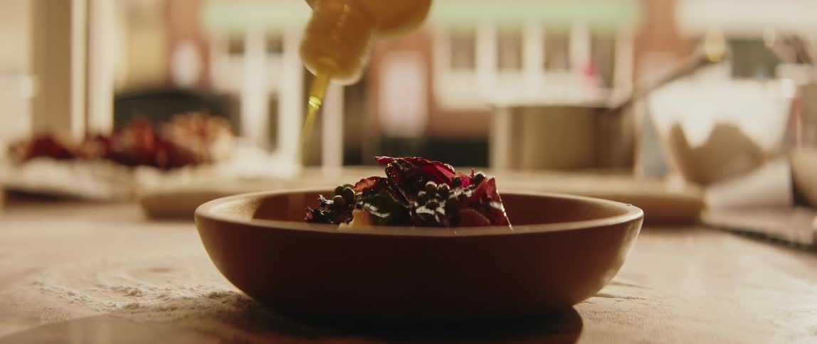 A chef pours golden oil over a dish of dark leafy greens and berries served in a shallow clay bowl on a flourdusted wooden table in a professional kitchen