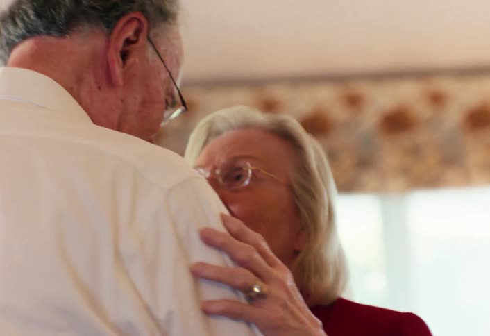 Elderly couple indoors with the woman resting her hand on the mans shoulder while they talk closely