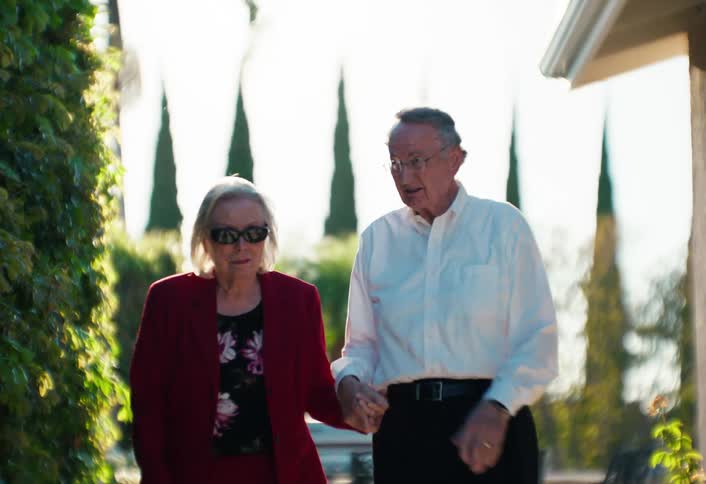 Elderly couple walking hand in hand in a residential outdoor area with the man gesturing while talking