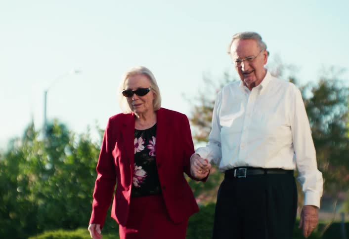 Elderly couple walking hand in hand outdoors with greenery and a clear sky in the background