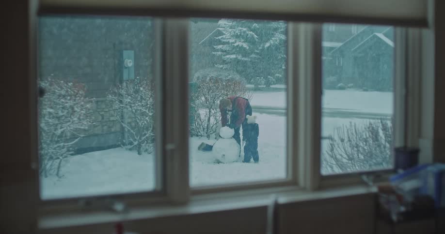 A man and two young children build a snowman in a snowy yard as seen through a window