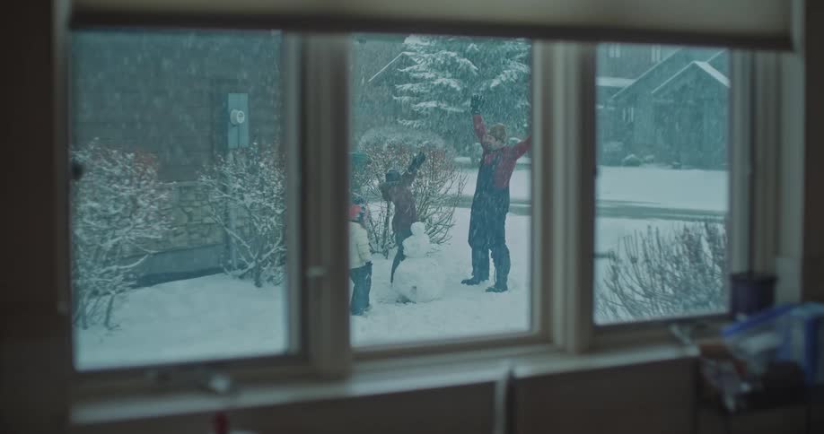 Children and an adult building a snowman in a yard during snowfall seen through a window