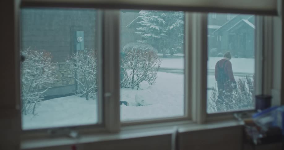 A father playing with three kids in the snow on a winter day building a snowman as the scene is viewed through a window