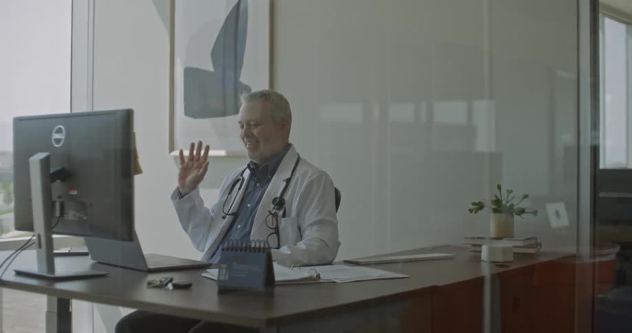 A doctor sits at his desk in a modern office smiling and waving during a video call on his computer