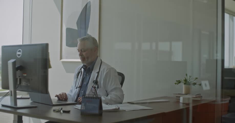 A doctor sits at a tidy desk in a modern office looking attentively at his computer with papers and a small plant nearby