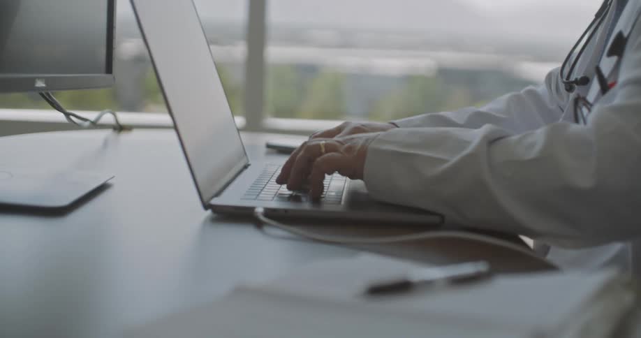 Doctor typing on a laptop at a work desk in a clinic with natural light in the background